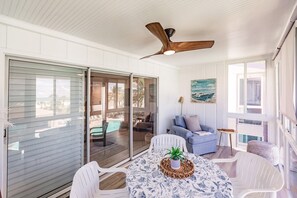 Um quarto iluminado pelo sol com uma porta de vidro deslizante para uma piscina, um ventilador de teto, uma mesa com uma planta e um quadro na parede.