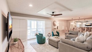 A spacious living room with a large TV, comfortable seating, a ceiling fan, and a view through the sliding glass doors. - 6018 - Gulfside Oasis with a beach view (Siesta Key)