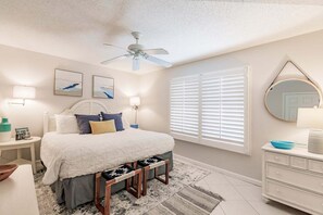 Quarto com cama, mesas de cabeceira, ventilador de teto, janela com persianas, cômoda e espelho.