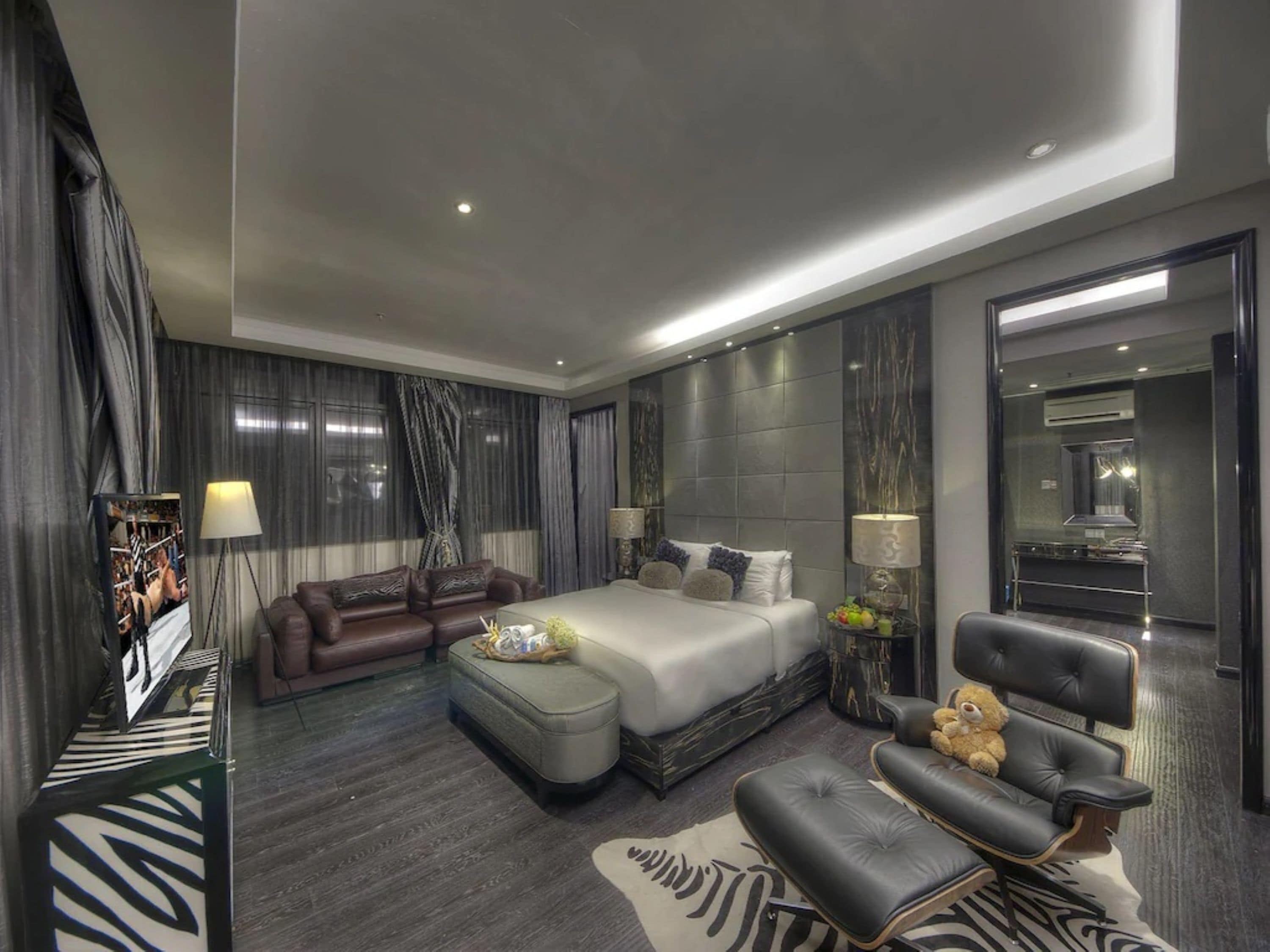A modern hotel room with a large bed, a sofa, a chair, and a small table with a teddy bear.