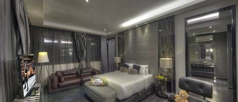 A modern hotel room with a large bed, a sofa, a chair, and a small table with a teddy bear.