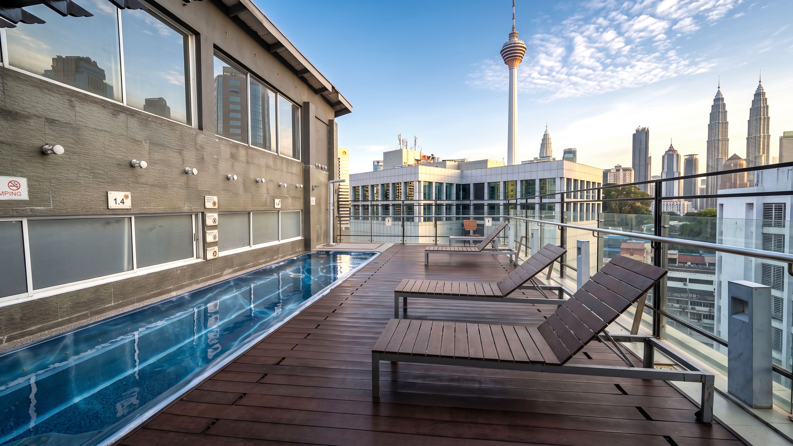 Área de piscina en la azotea con sillones de descanso, barandilla de vidrio y vista al horizonte urbano que incluye una torre.