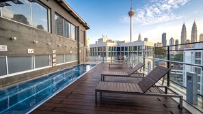 Área de piscina en la azotea con sillones de descanso, barandilla de vidrio y vista al horizonte urbano que incluye una torre.