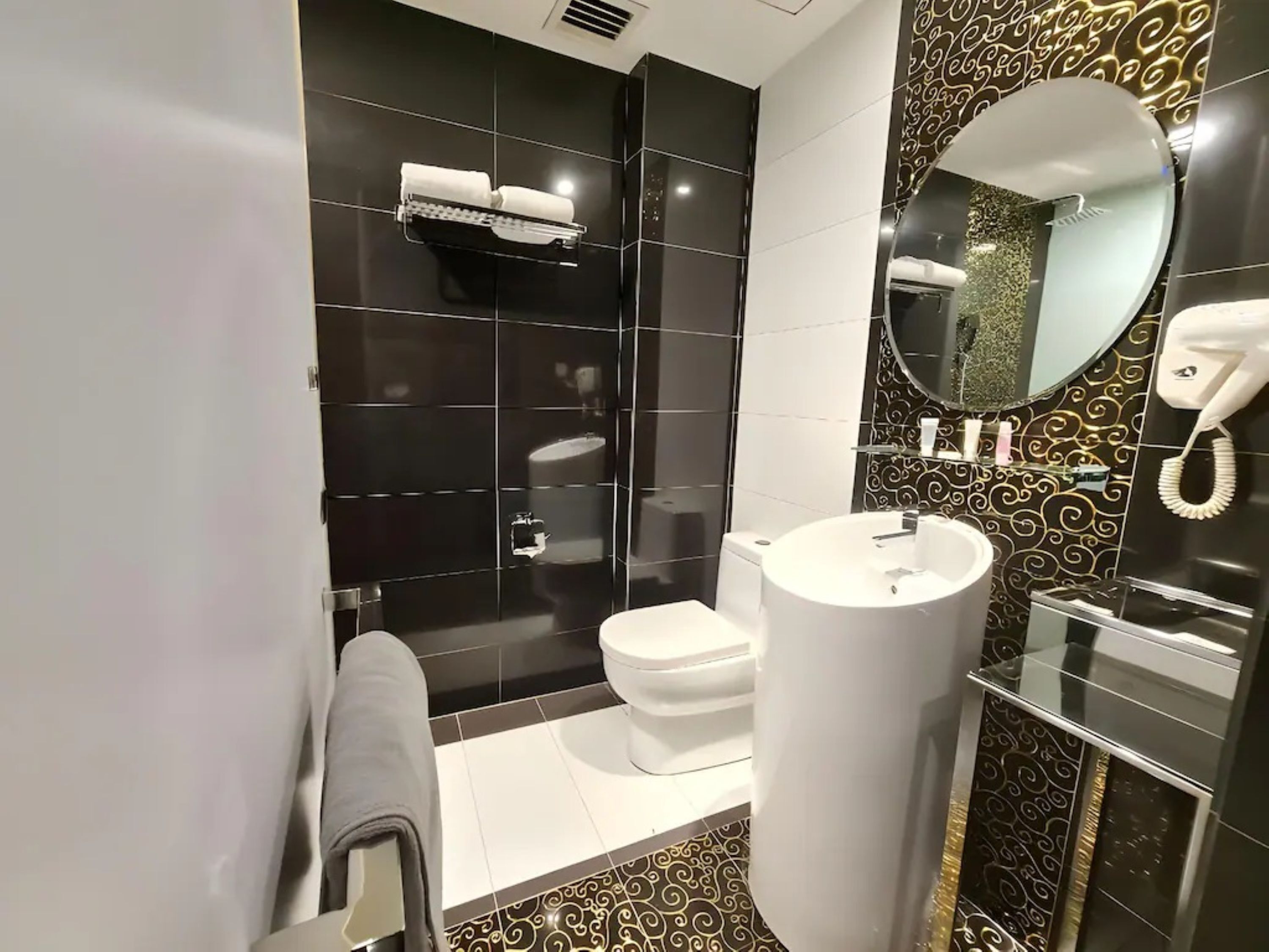A modern bathroom with a white sink, toilet, and shower area. There is a towel rack and a mirror with a telephone on the wall.