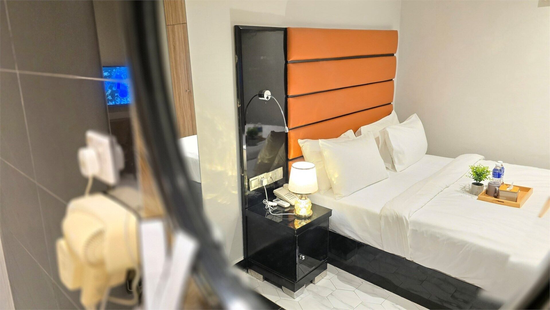 Habitación de hotel con cama, mesita de noche, lámpara y un espejo que refleja parte del cuarto.