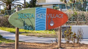 A sign for Sierra Key Village with a natural design on one side and a modern building in the background. - SR-12 Gulf Front 2/1 (Siesta Key)