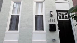 A multi-story building with a white facade, numbered 1415, featuring multiple windows with white frames and curtains, a black door, and a small mailbox.