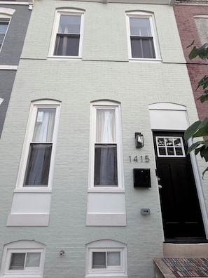 A multi-story building with a white facade, numbered 1415, featuring multiple windows with white frames and curtains, a black door, and a small mailbox.