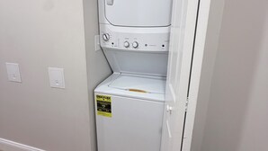 A white laundry room with a built-in washer and dryer, a small shelf with cleaning supplies, and a power outlet on the wall.