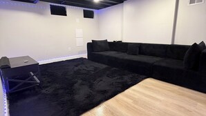 A room with a black sofa, a wooden table, and a black rug. There are ceiling lights and a white wall with a small window.