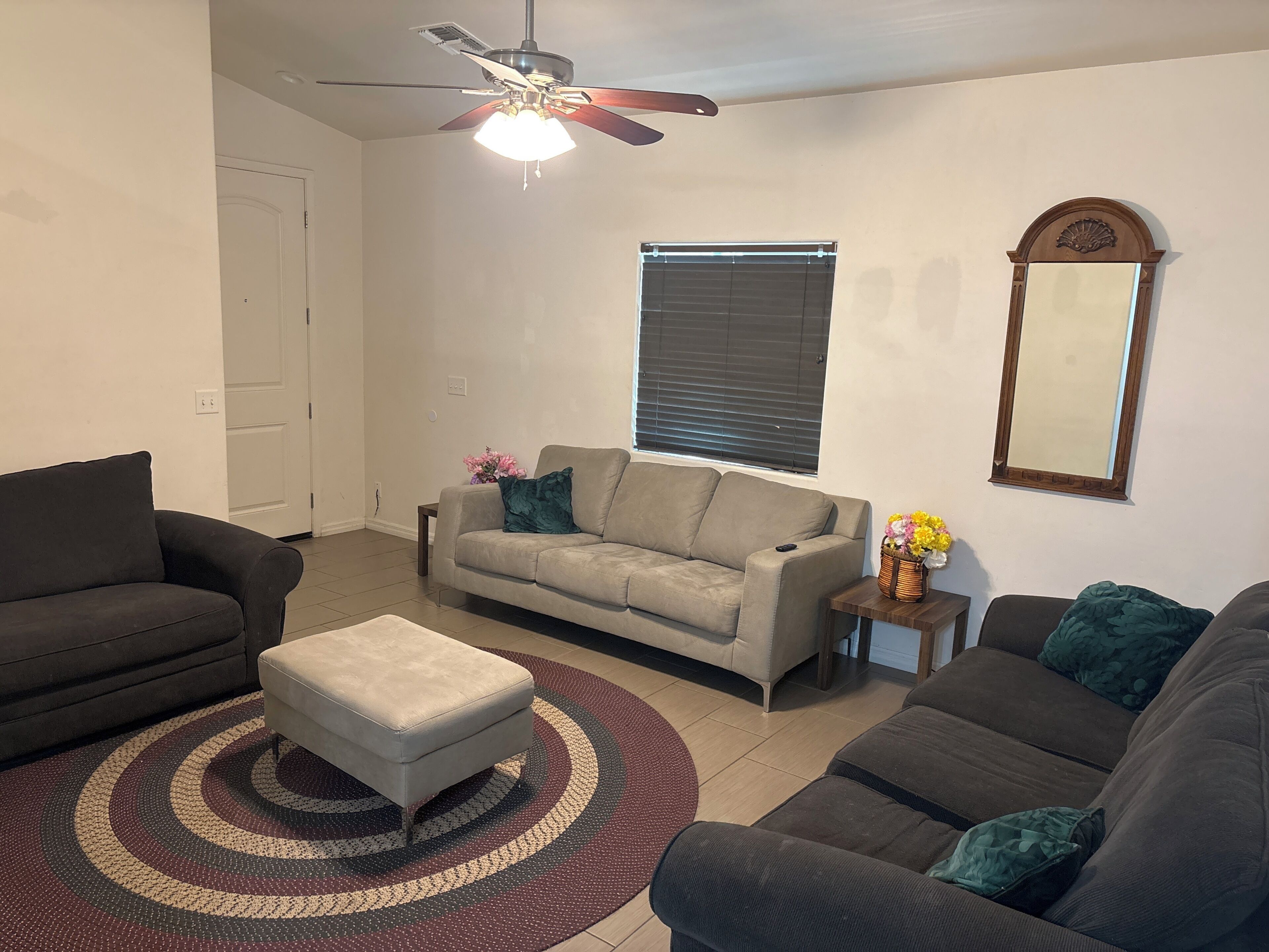 A living room with a ceiling fan, a sofa, an ottoman, a small table with a vase, and a window with blinds.