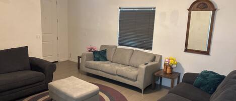 A living room with a ceiling fan, a sofa, an ottoman, a small table with a vase, and a window with blinds.