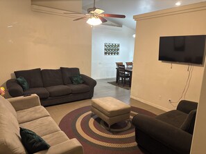 Ein Wohnzimmer mit braunem Sofa, einem beigefarbenen Sessel, einem gemusterten Teppich, einem Deckenventilator und einem an der Wand befestigten Flachbildfernseher.