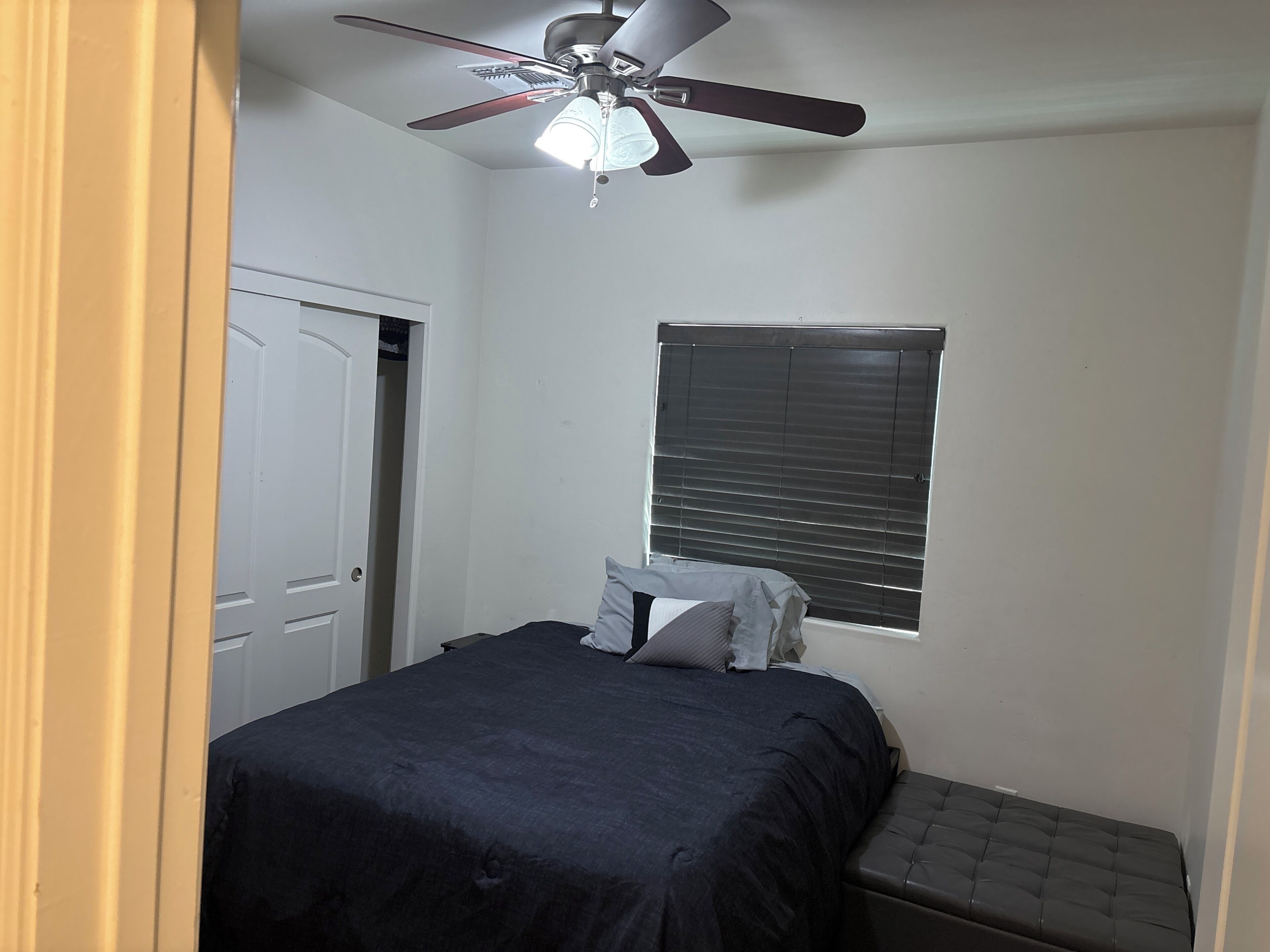 Ein Schlafzimmer mit einem Bett, einem Deckenventilator, einem Fenster mit Jalousien und einer Tür.