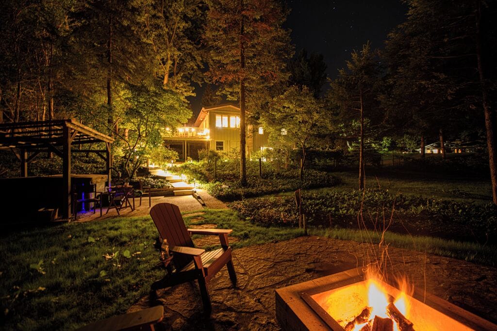 A nighttime outdoor setting with a fire pit, seating area, and a house with illuminated windows.