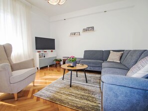 Ein modernes Wohnzimmer mit grauem Sofa, einem hellen Sessel, einem Couchtisch und einem an der Wand befestigten Flachbildfernseher.