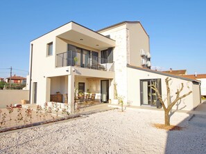 Um edifício residencial de dois andares com varanda, área de estar ao ar livre e entrada por um caminho de cascalho.