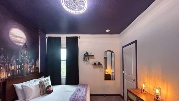 Ein Schlafzimmer mit einem Bett, einem Schminktisch, einem Spiegel, einem Fenster mit Vorhängen und einem Wandbild.