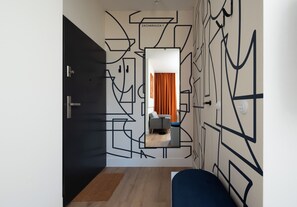 A hallway with a geometric wall design, a mirror reflecting a room with a sofa and curtains, and a door with a handle. - Zachariasza Harmony Apartment (Warszawa)