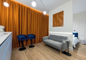Habitación de hotel moderna con sofá gris, dos sillas azules, mesita de noche blanca y suelo de madera.
