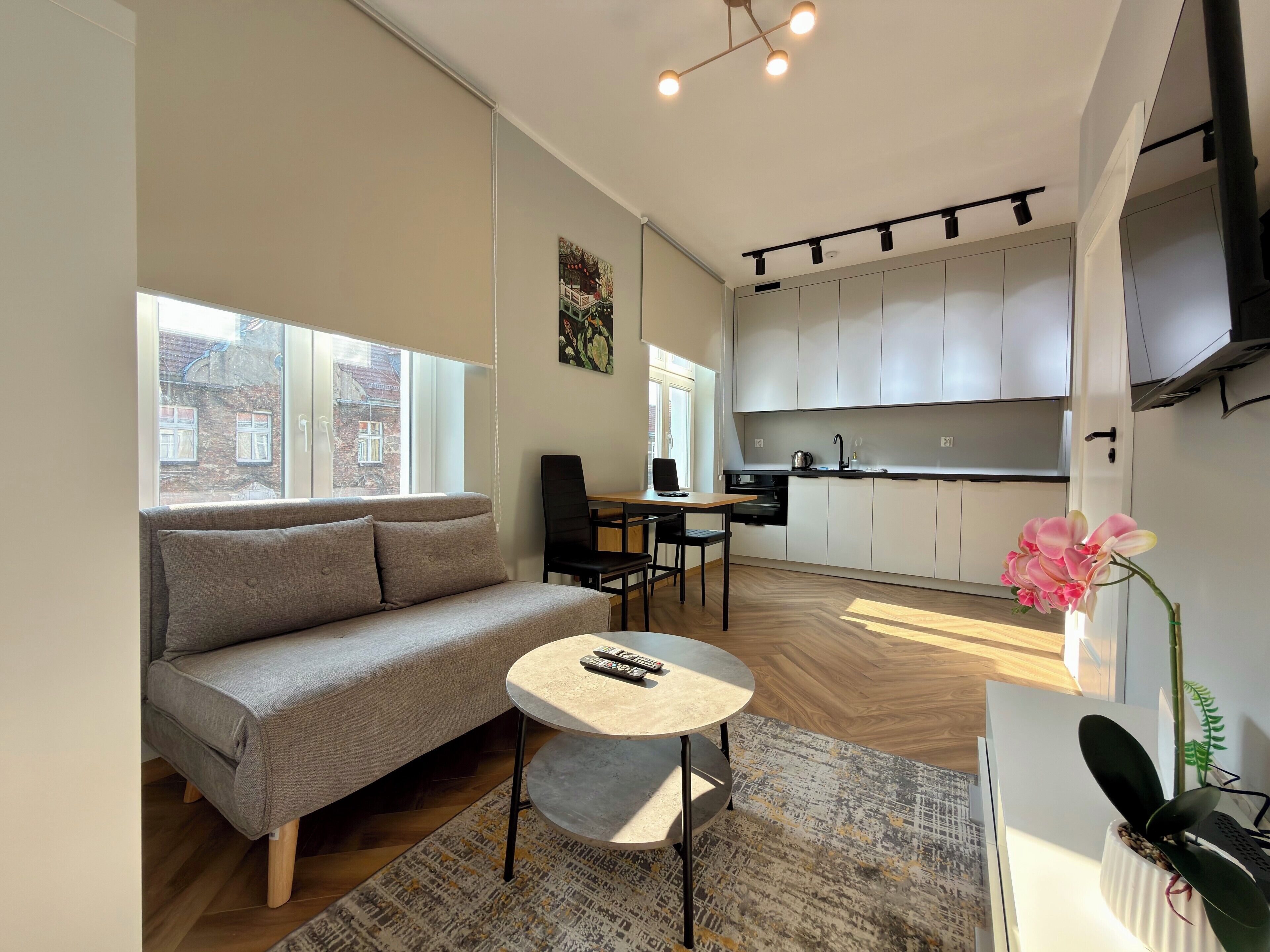 A modern living space with a sofa, a small round table, a chair, a kitchen area with cabinets, and a window with a view of buildings.