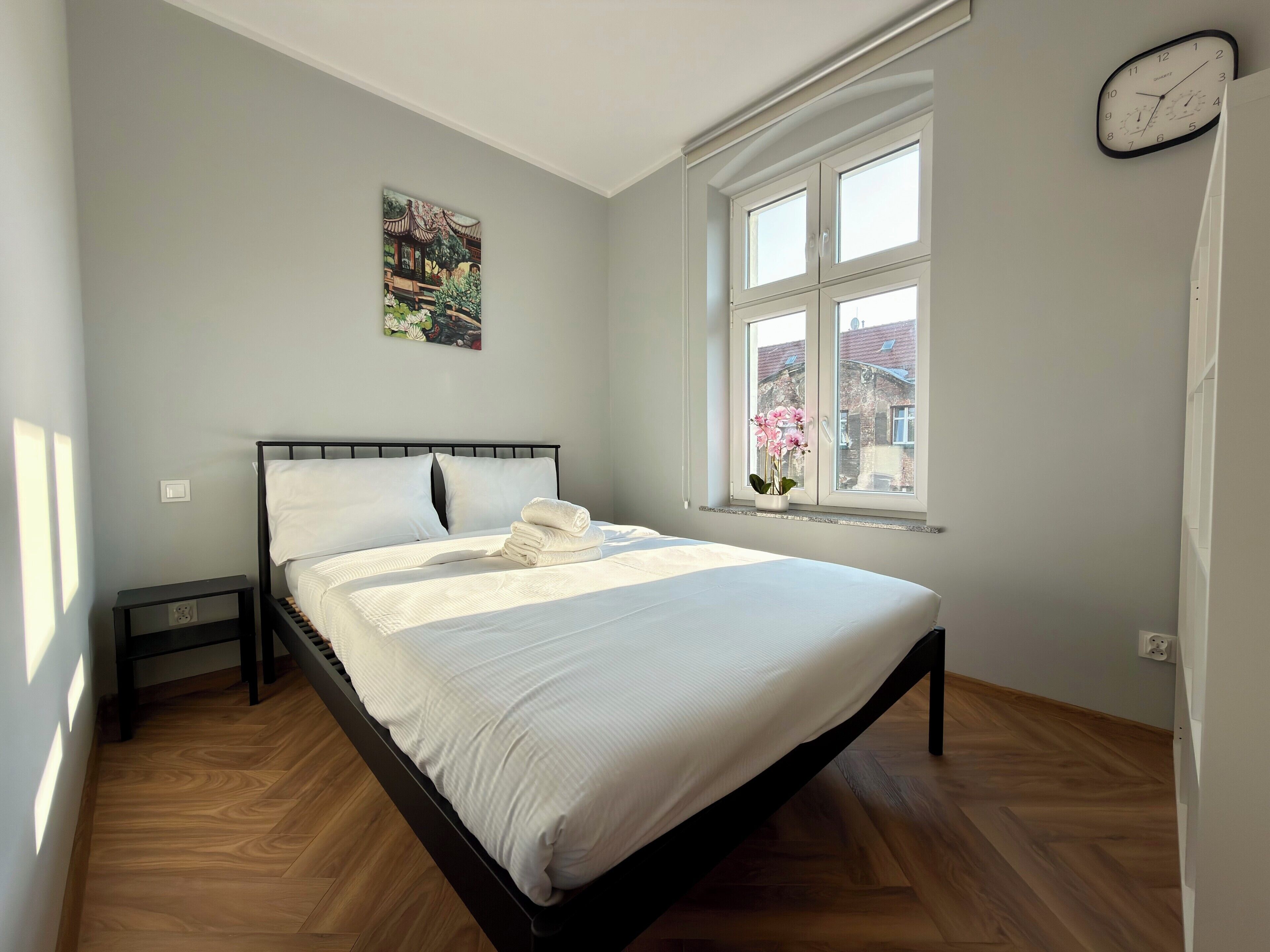 Un dormitorio con cama, mesita de noche, ventana con vistas y un reloj de pared.