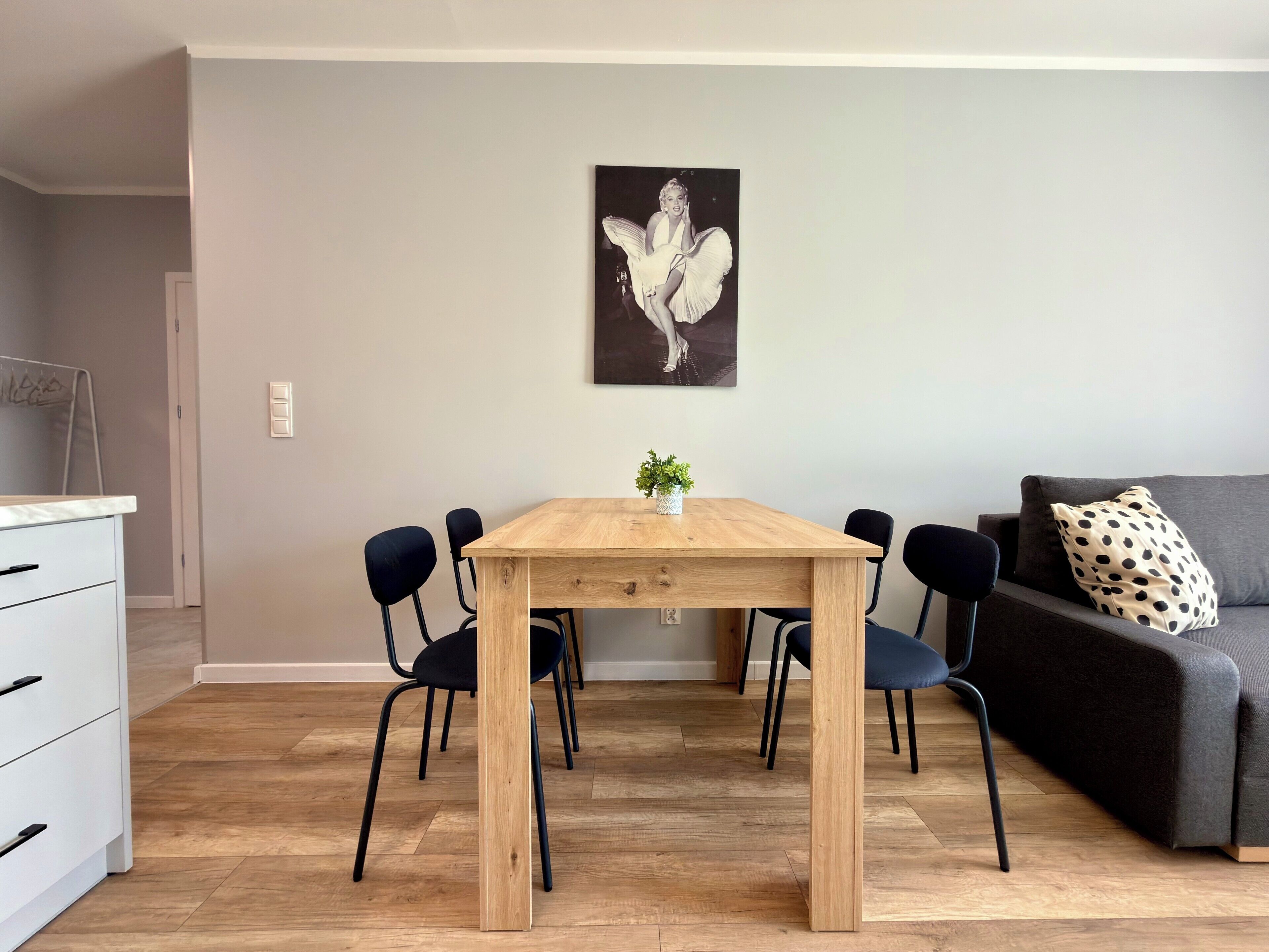 A dining area with a wooden table, four chairs, a small plant, and a grey sofa with a polka-dotted pillow.
