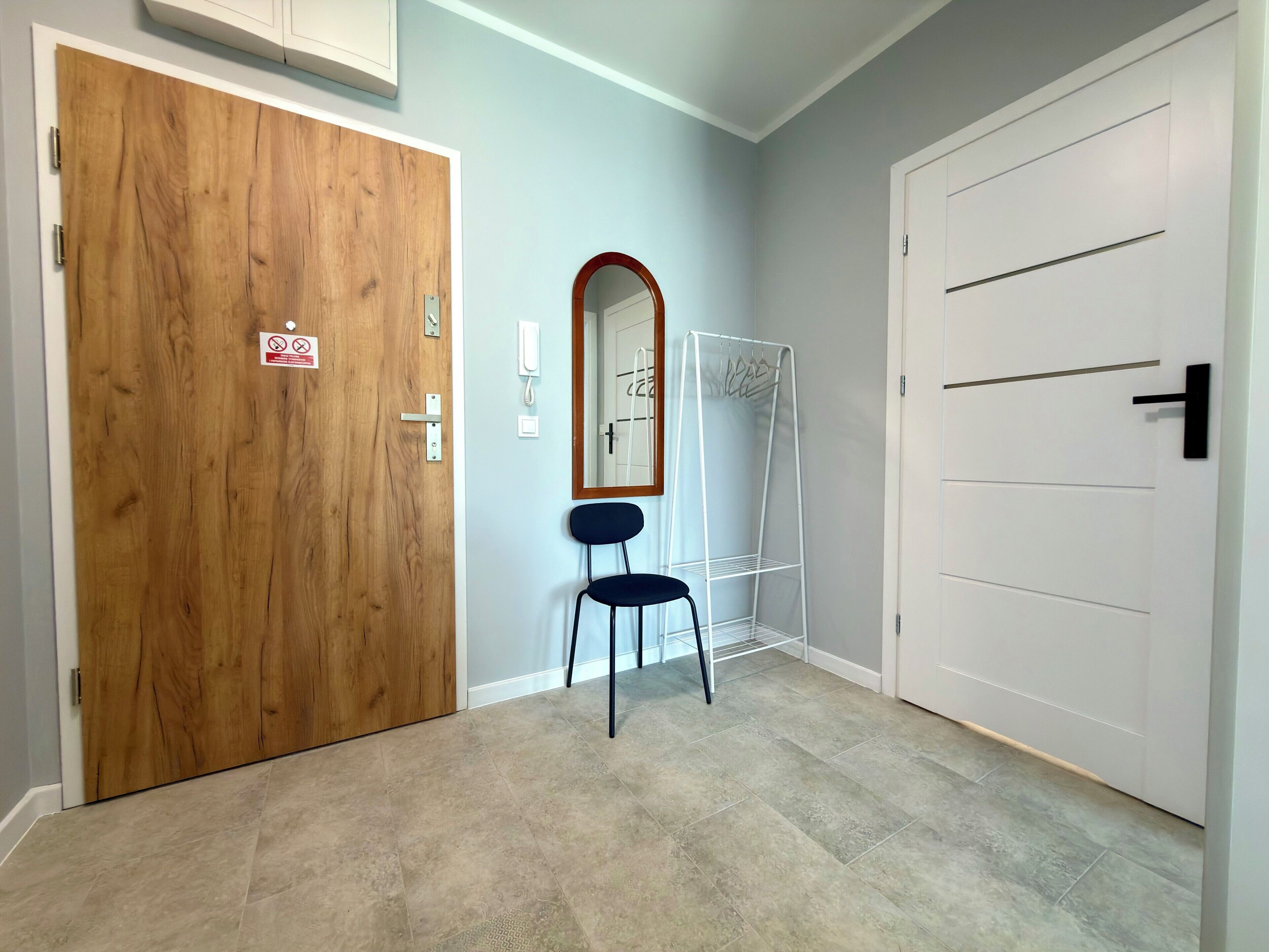 A hallway with a wooden door, a white door, a mirror, a metal drying rack, and a black chair.