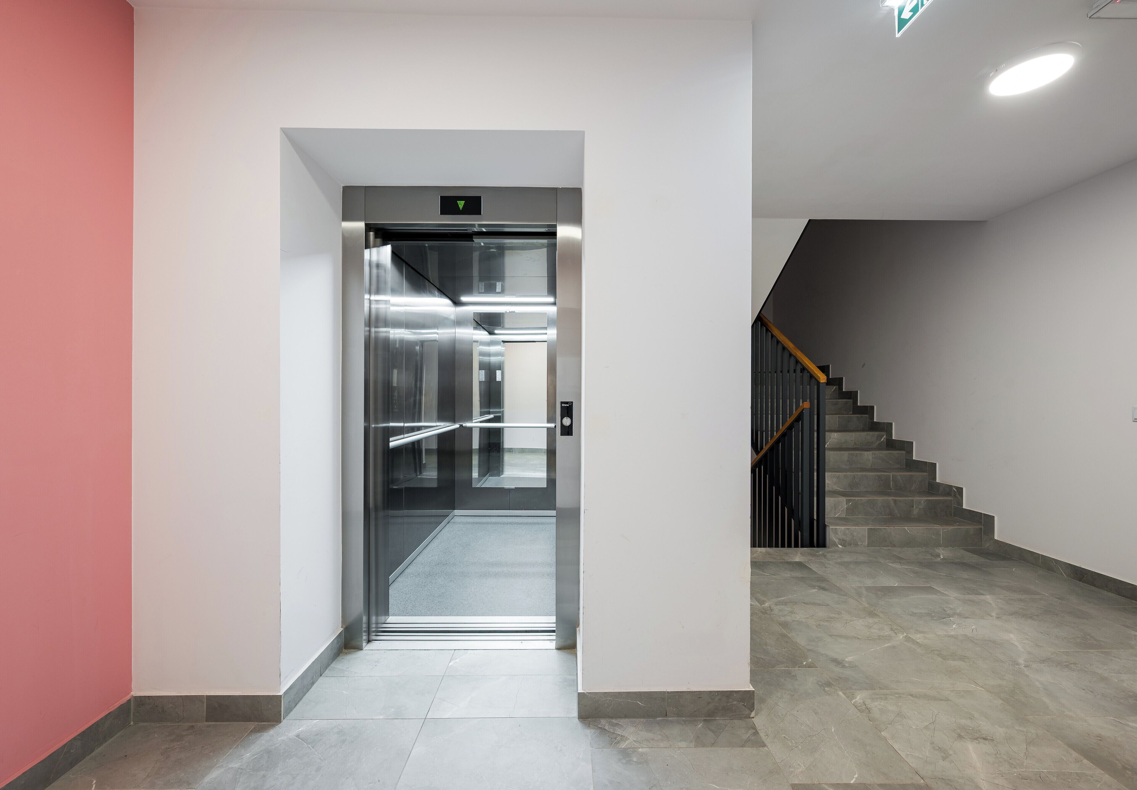 Ein Aufzug mit metallischem Innenraum und Handlauf, der zu einer Treppe mit Holzwurf und grauen Fliesen führt.