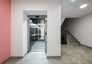 An elevator with a metallic interior and a handrail, leading to a staircase with a wooden handrail and grey tiles. - Kłobucka Harmony Apartment (Warszawa)