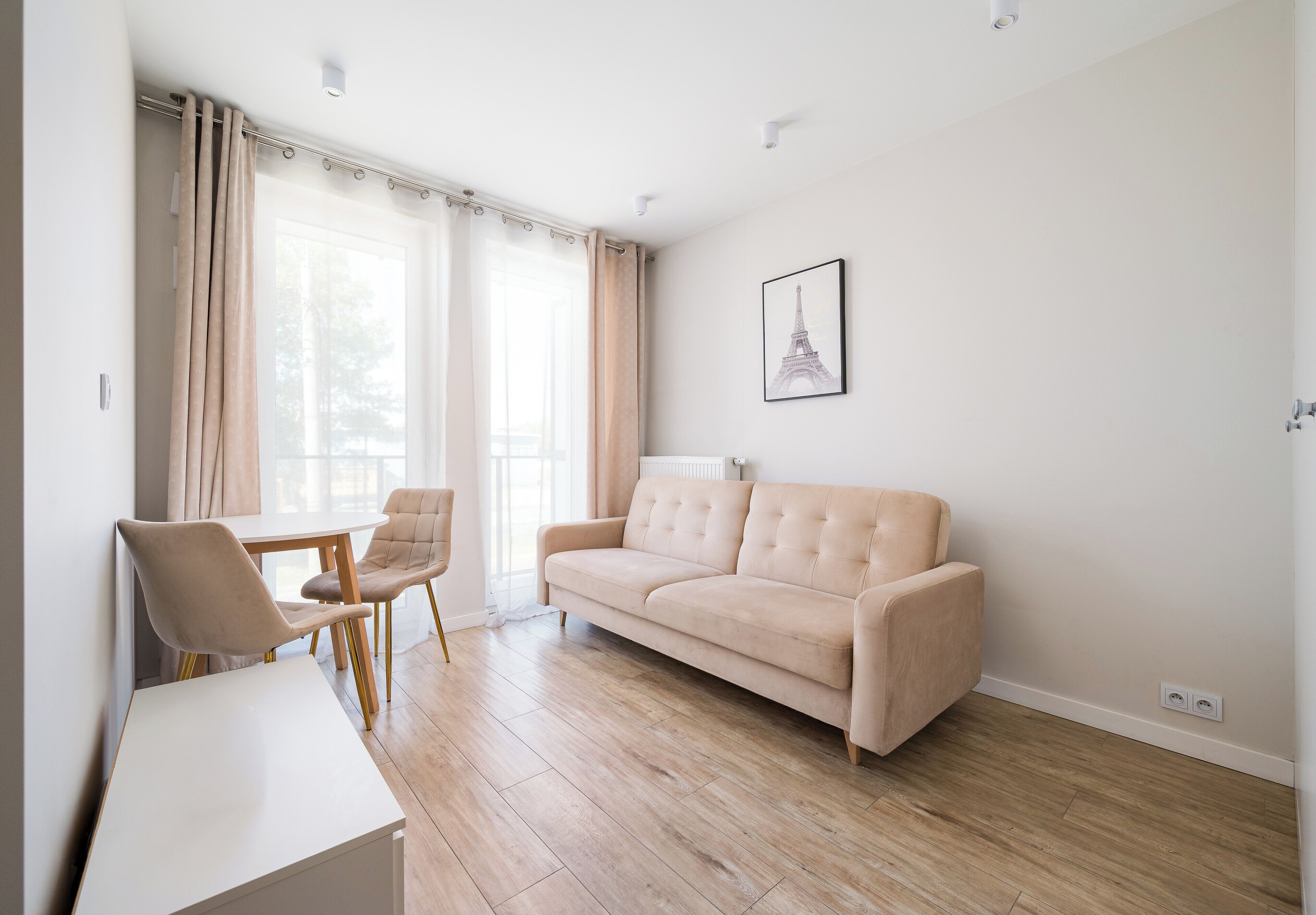Ein modernes Wohnzimmer mit einem beigefarbenen Sofa, einem Esstisch mit zwei Stühlen und einem großen Fenster mit Vorhängen.
