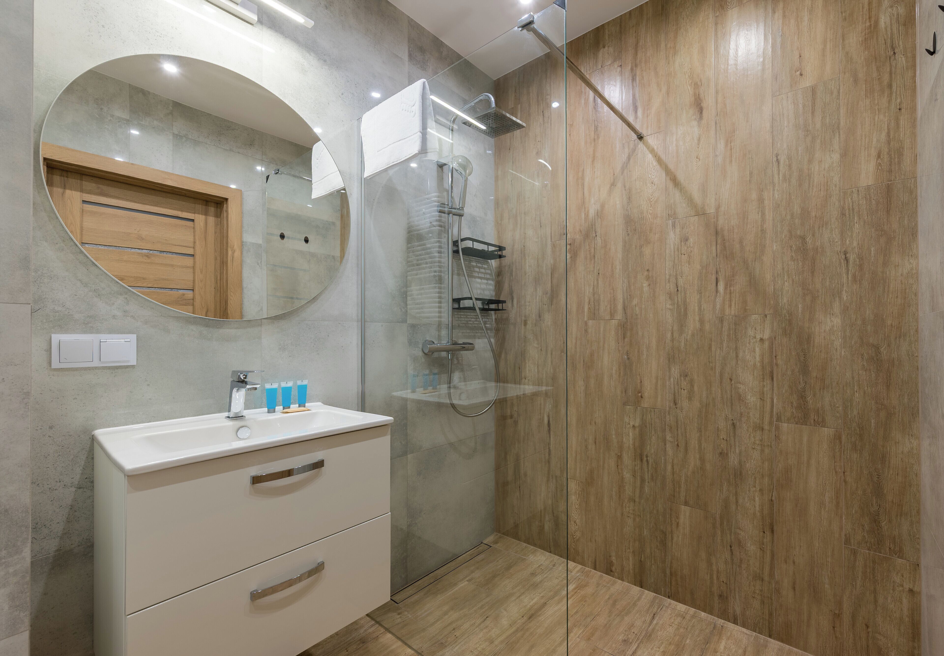 A modern bathroom with a glass-enclosed shower, a round mirror, and a wooden vanity.