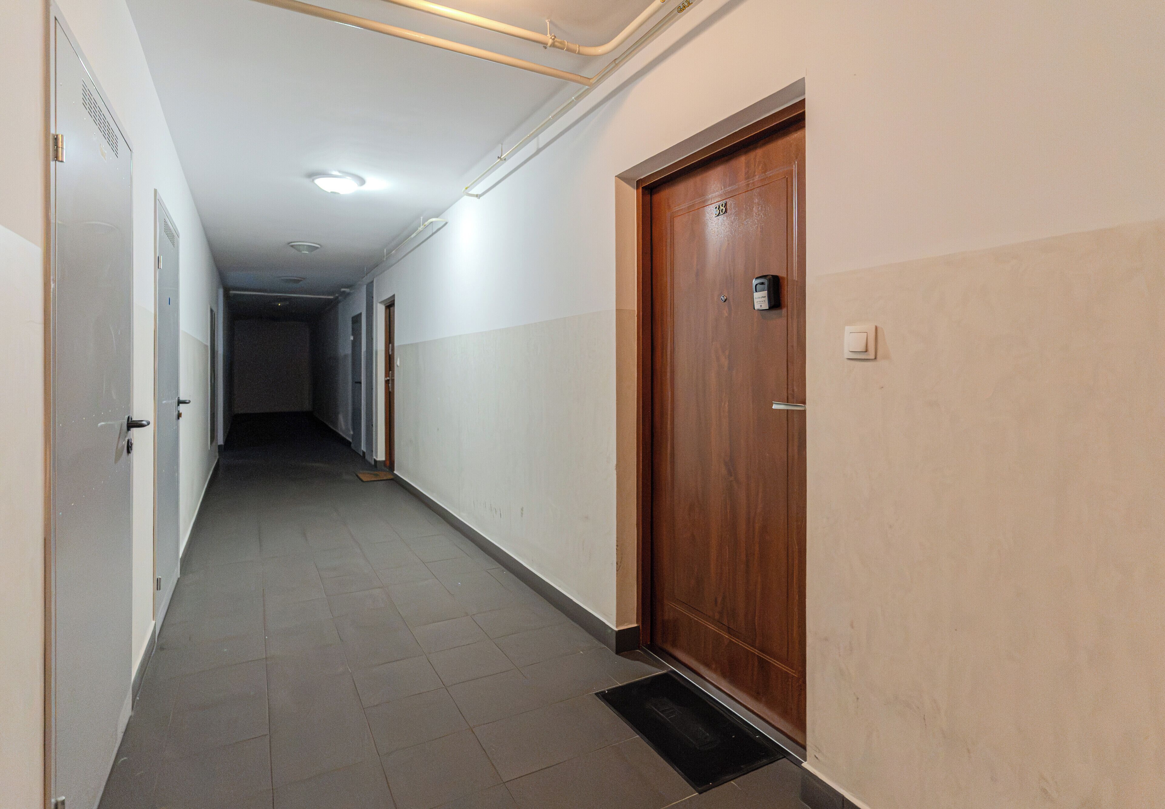 Um corredor longo e estreito com uma porta de madeira à direita, uma porta de metal à esquerda e uma abertura no teto.