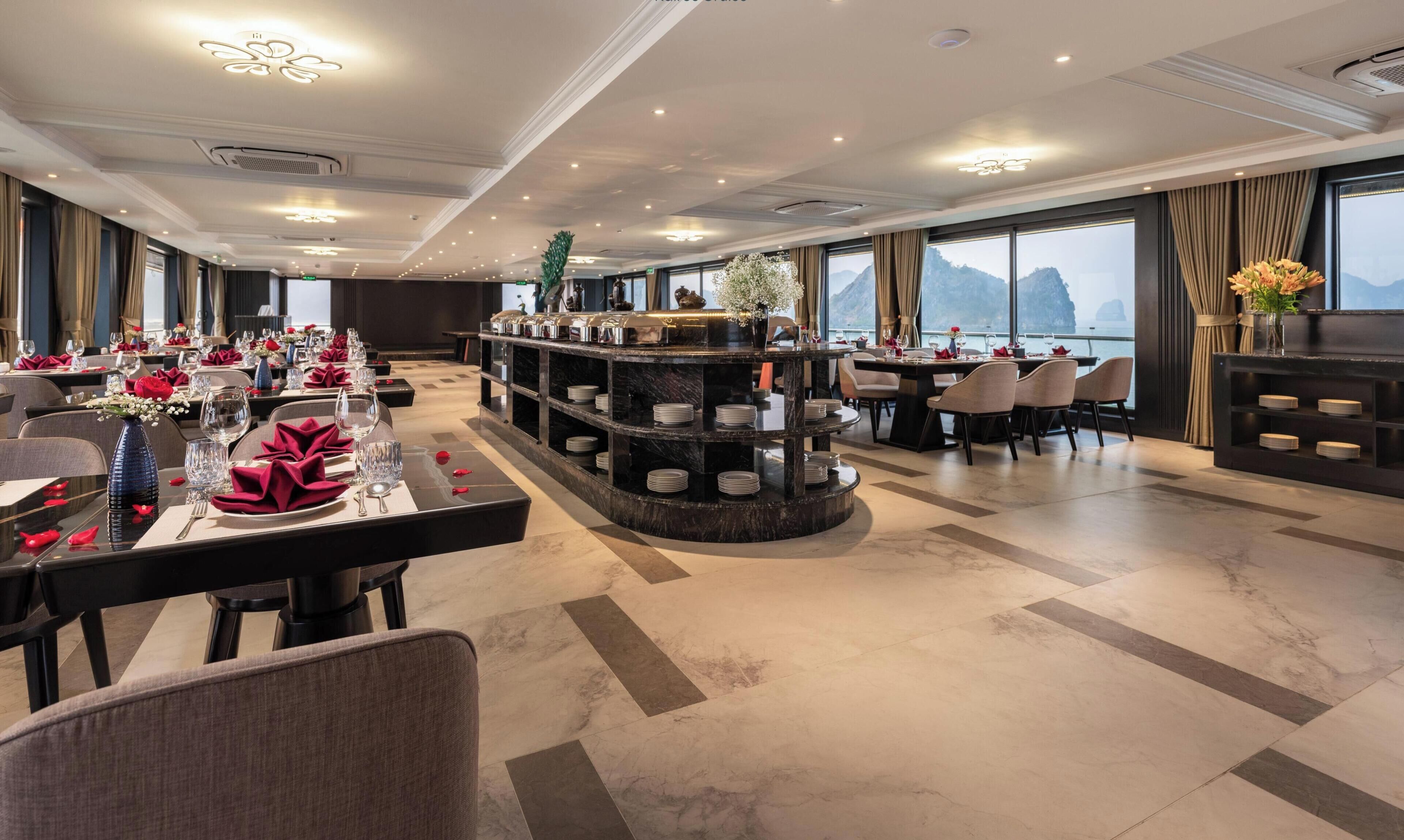 A hotel dining area with multiple tables set for guests, a central bar area with shelves, and large windows offering a view of the mountains.