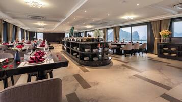A hotel dining area with multiple tables set for guests, a central bar area with shelves, and large windows offering a view of the mountains.