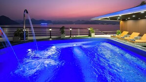 Ein Swimmingpool mit Wasserfall, umgeben von Liegestühlen und Topfpflanzen, mit Blick auf ein Gewässer und Berge in der Abenddämmerung.