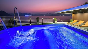 Piscina con cascada, rodeada de sillas de descanso y plantas en macetas, con vistas a un cuerpo de agua y montañas al anochecer.