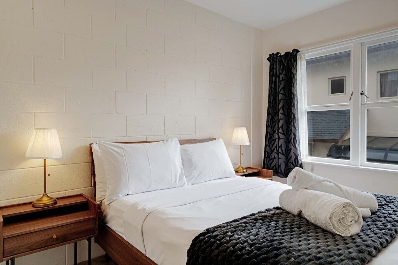 A hotel room with a bed, two bedside tables, lamps, a window with curtains, and a view of the outside.