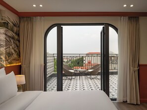 Habitación de hotel con cama, mesita de noche, lámpara, vista de la ciudad a través de una puerta de vidrio y balcón con mesa y sillas.