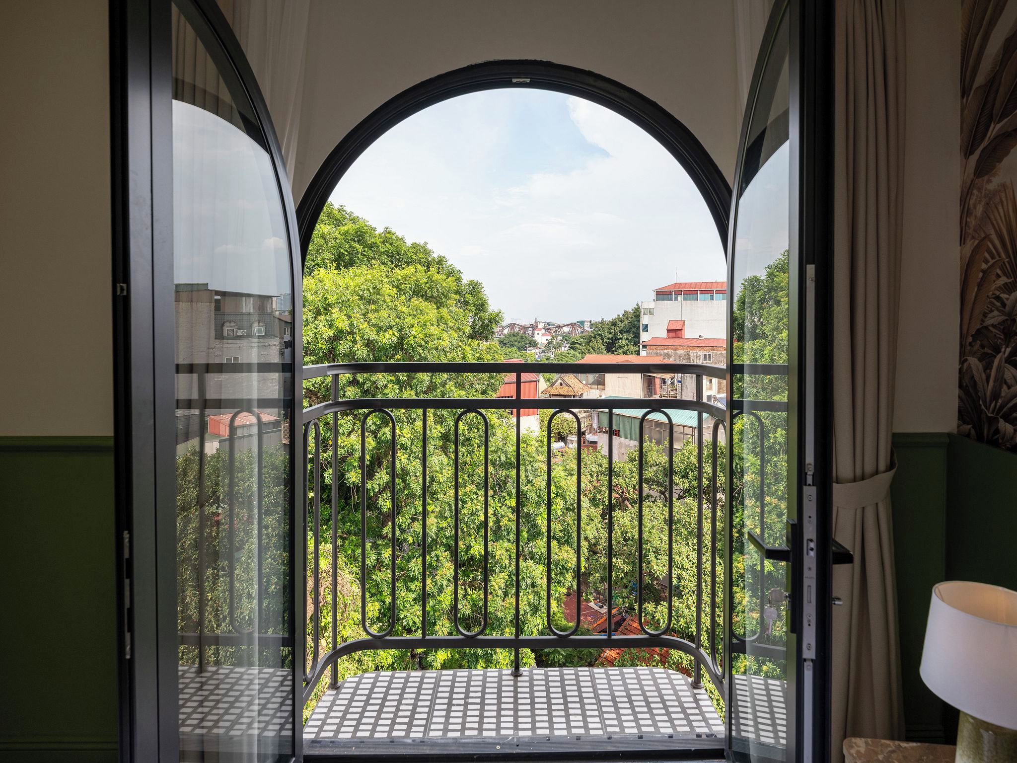 A balcony with a metal railing, a view of greenery and buildings, and a partial view of a room with a lamp.
