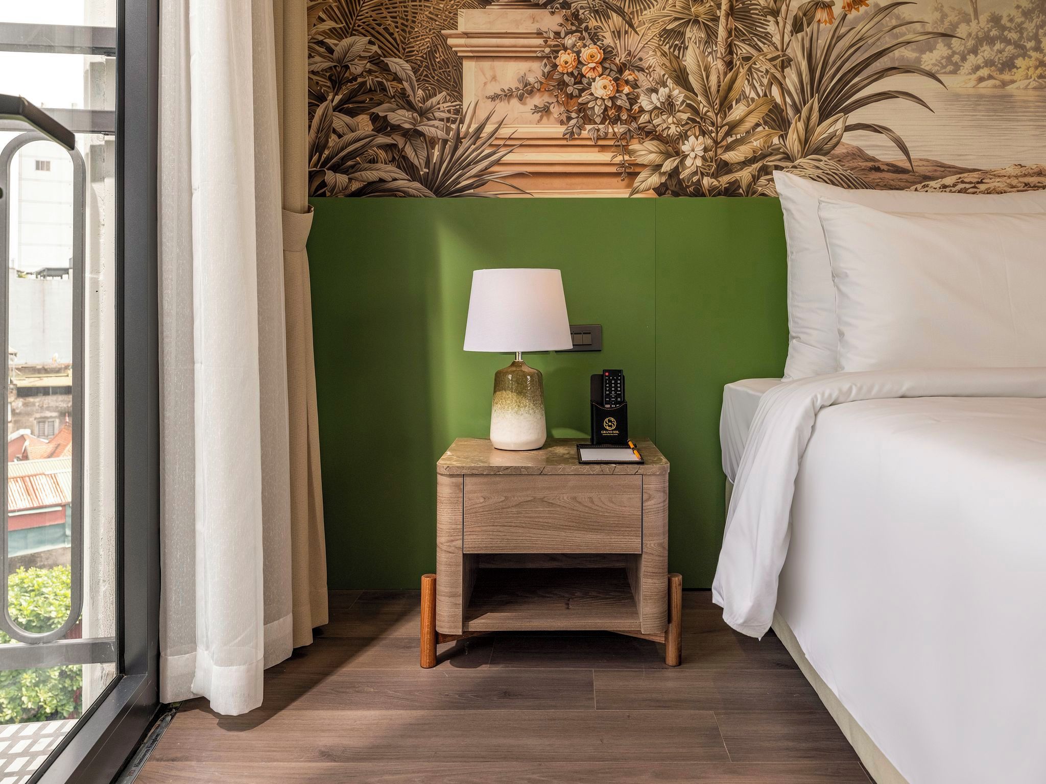A hotel room with a bed, a nightstand with a lamp, a remote control, and a wall with a floral mural.