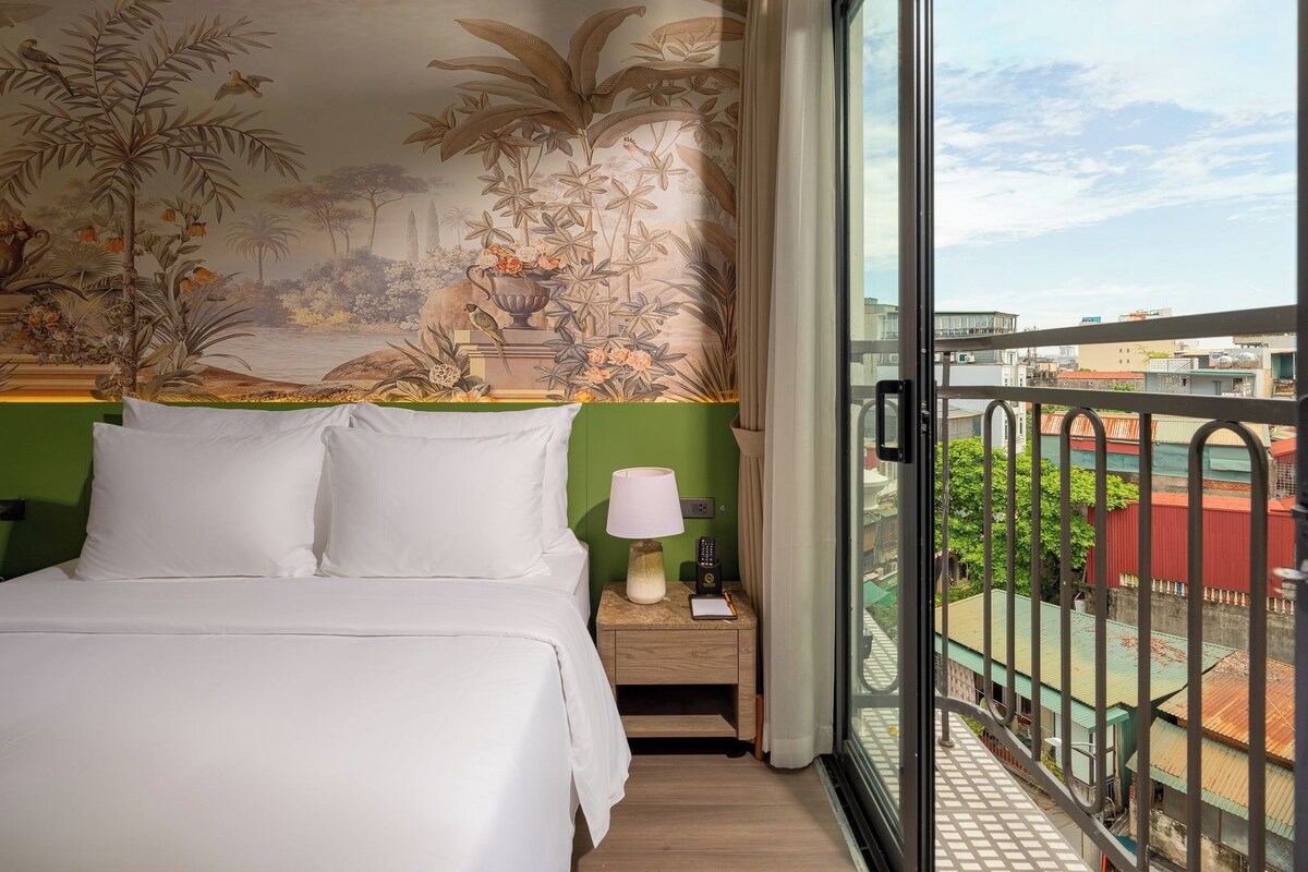Habitación de hotel con cama, mesita de noche, lámpara y vistas a la ciudad desde un balcón.