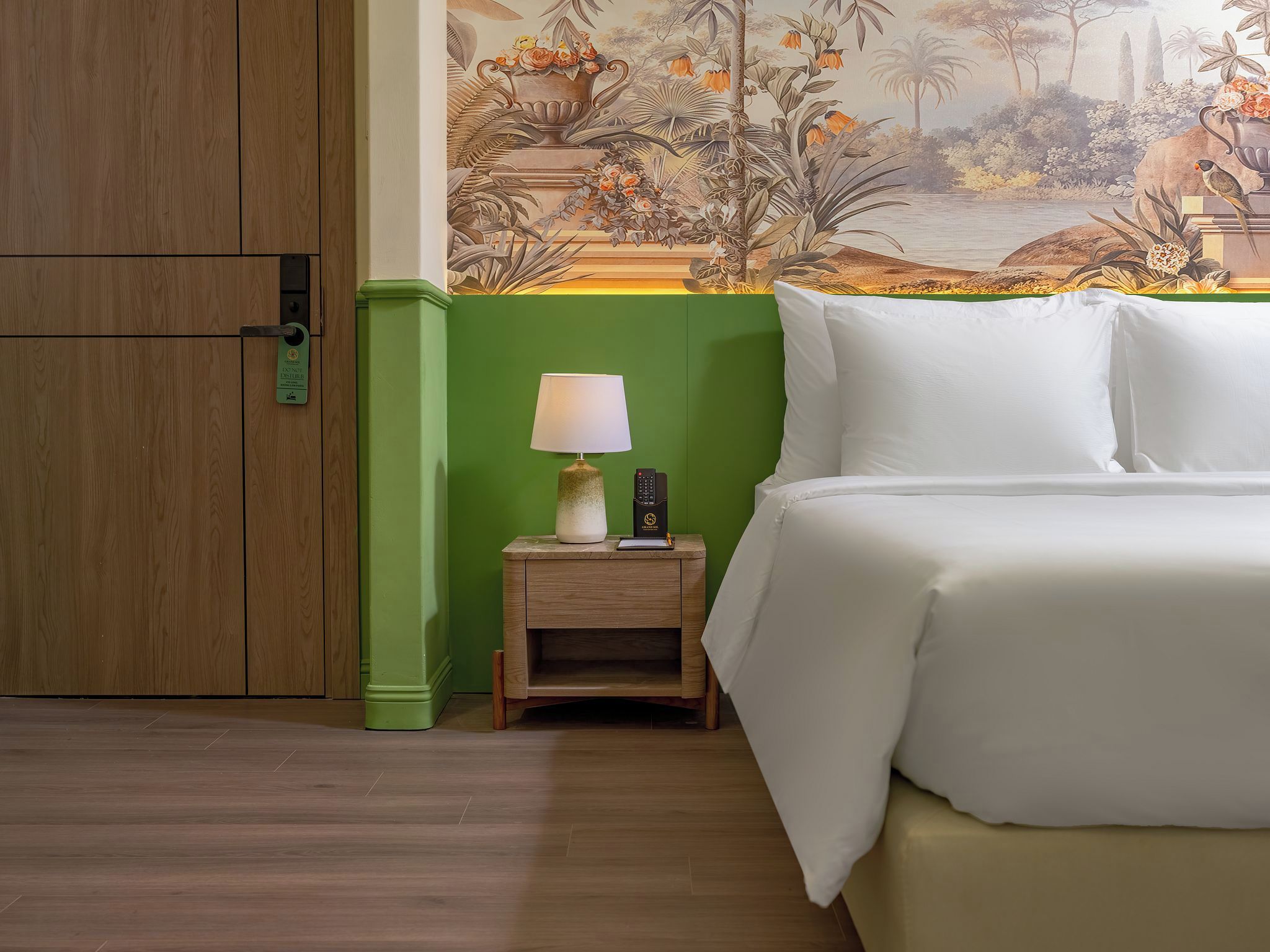 Een hotelkamer met een bed, een nachtkastje met een lamp, een groene wand en een muurschildering van een tropisch landschap.