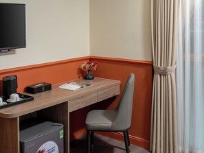 Habitación de hotel con un escritorio de madera, una silla, un televisor montado en la pared, una mesita con un florero y una ventana con cortinas.