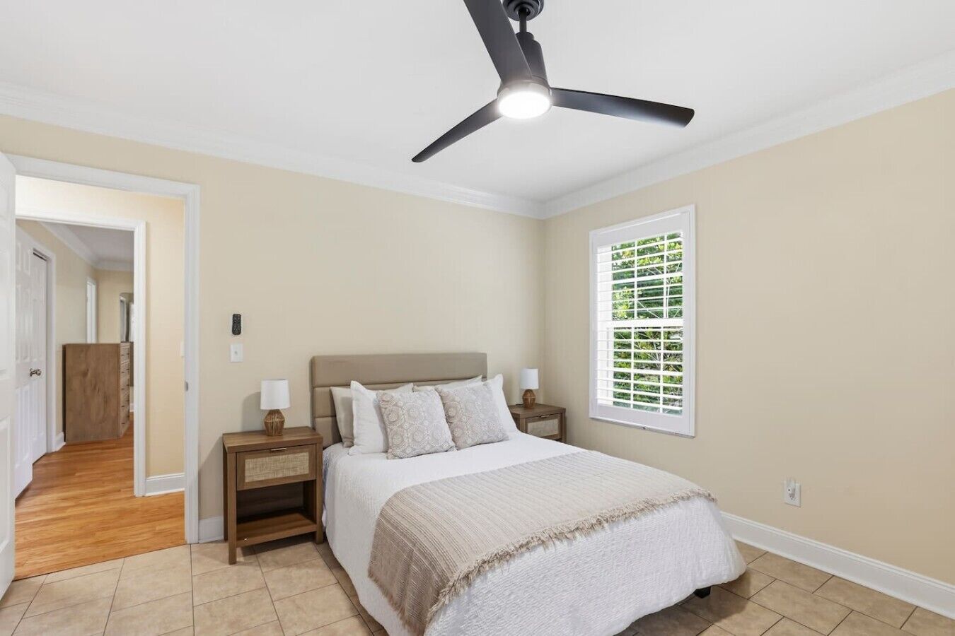 A bedroom with a bed, nightstands, a ceiling fan, a window with blinds, and a door leading to another room.