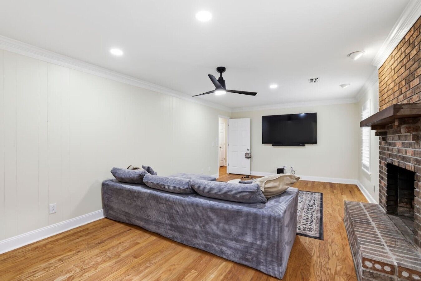 A modern living room with a grey sofa, a flat-screen TV mounted on the wall, a ceiling fan, and a brick fireplace.
