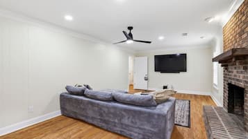A modern living room with a grey sofa, a flat-screen TV mounted on the wall, a ceiling fan, and a brick fireplace.