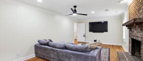 A modern living room with a grey sofa, a flat-screen TV mounted on the wall, a ceiling fan, and a brick fireplace.