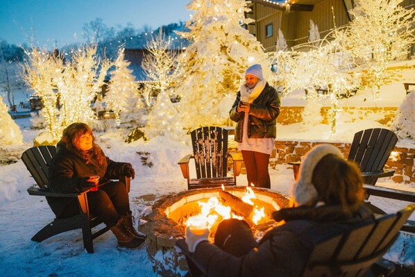 Um grupo de pessoas reunido em volta de uma fogueira em ambiente externo coberto de neve, com árvores carregadas de neve e um edifício ao fundo.