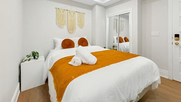 Een netjes opgemaakt bed met een wit-oranje dekbedovertrek, een klein nachtkastje met een plantje en een spiegel aan de muur.